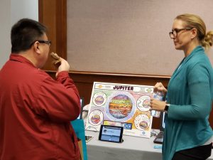 Woman standing at a table talking with a man. On the table is a small tablet and a large poster with facts about Jupiter.